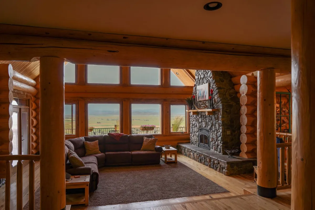 A spacious living room with a large, traditional stone fireplace set against a wall of expansive windows, allowing sweeping mountain views. A comfortable sectional sofa is arranged for cozy gatherings, and the room is bathed in warm, welcoming light.