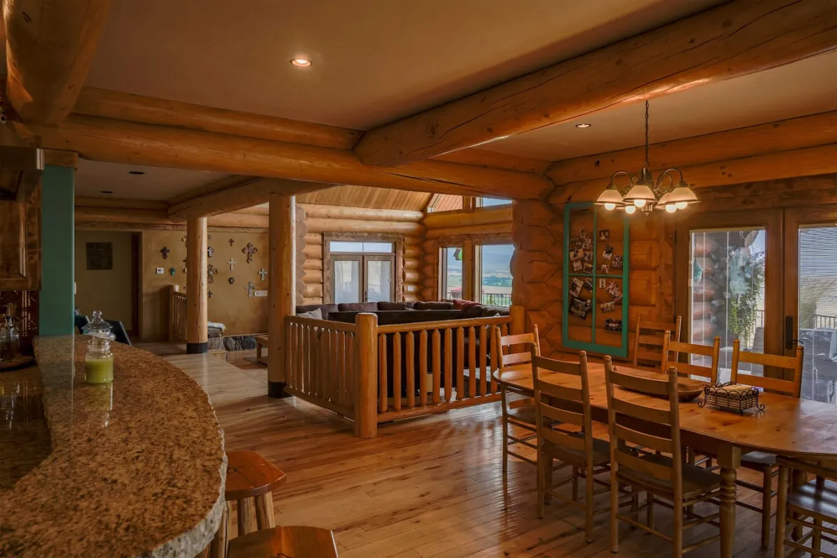 An inviting open-concept dining area featuring a large wooden dining table that can seat a group, a granite bar counter for casual meals or celebratory toasts, and log cabin walls. Large windows provide more views and natural light, reinforcing the communal atmosphere.
