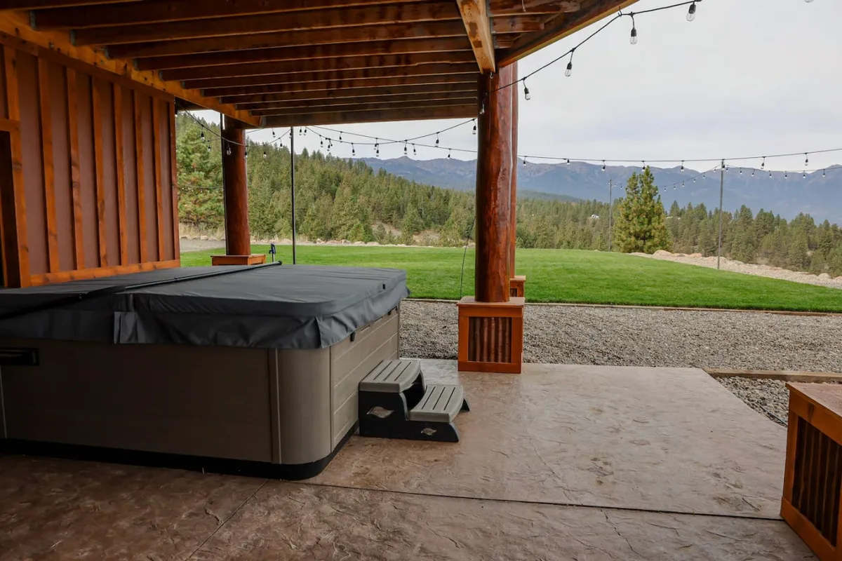 Set against a backdrop of lush green lawn and mountain vistas, a hot tub framed by wooden columns promises relaxation and privacy. The scene suggests soaking under the stars, sharing conversations, and enjoying the serenity of nature—all key aspects of a romantic or restful escape.