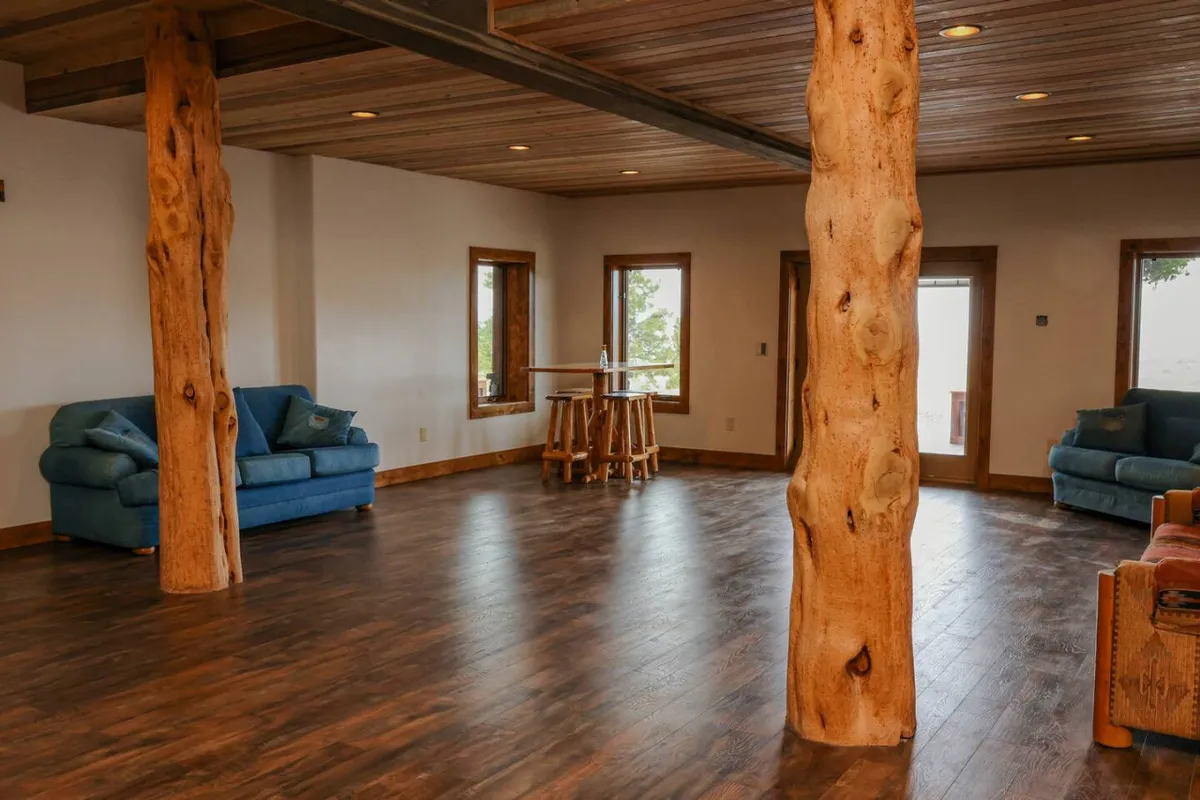 A stylish, open-plan living area with blue sofas, sturdy wooden pillars, and a bar table with stools. Large windows provide scenic views and create a bright, airy ambiance. The furniture is arranged to foster social interaction and relaxation, making it perfect for unwinding after activities or gathering for after-adventure refreshments.