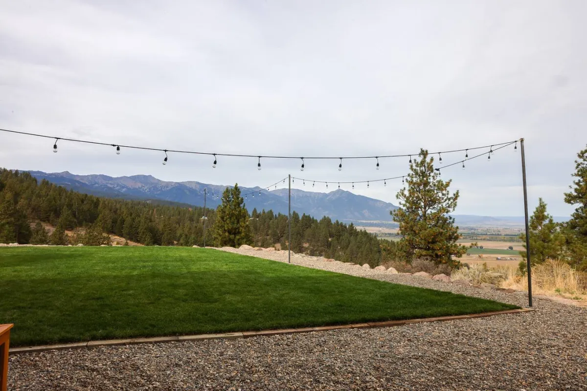 A beautifully landscaped lawn stretches out beneath the glow of string lights, with pine trees and distant mountain views providing a serene natural backdrop. The combination of the expansive green space and gentle lighting creates a romantic atmosphere perfect for evening gatherings, stargazing, or quiet moments with loved ones.