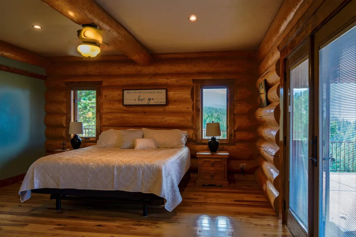 A tranquil bedroom featuring log walls, a bed adorned with decorative pillows, nightstands with lamps, and large windows revealing scenic mountain views. Natural light streams in, creating a peaceful retreat ideal for relaxation and restful sleep.