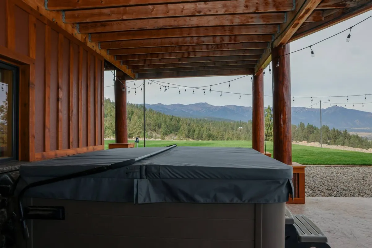 An exterior view of the grand deck featuring a bubbling hot tub beneath string lights, with wooden beams and a breathtaking mountain vista in the background. The scene radiates tranquility and invites guests to soak, unwind, and take in sunsets or stargaze after a day of explorations.