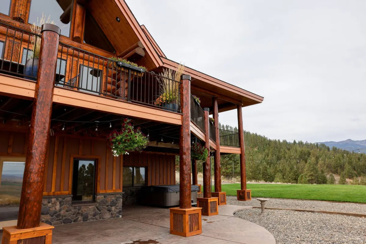 This exterior shot features the lodge's impressive wooden columns, a stone facade, a balcony adorned with flowers, and a sprawling green lawn that stretches out before a dramatic mountain backdrop. The setting is open, welcoming, and clearly designed to accommodate large groups for gatherings.