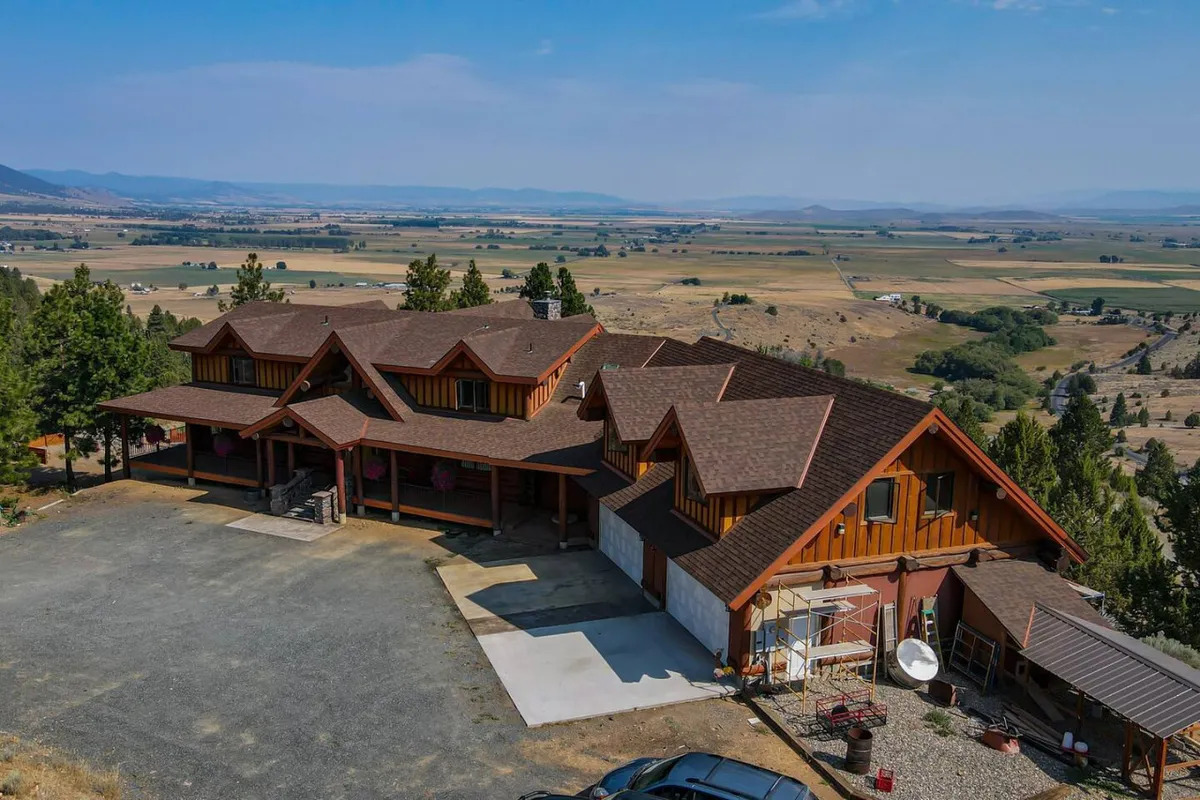 A panoramic exterior view of a spacious, multi-level house surrounded by natural landscape and mountains. The ample parking area and outdoor spaces suggest easy access for multiple vehicles and room for large gatherings, such as reunions or corporate events.