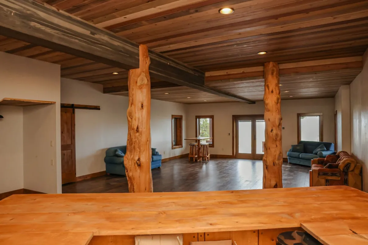 A spacious, inviting basement living room with prominent wooden pillars, large windows letting in natural light, an open layout, a substantial wooden bar counter, and multiple seating areas perfect for group gatherings or casual relaxation. The scene conveys a comfortable, multi-purpose space ideal for socializing, movie nights, or playing games.