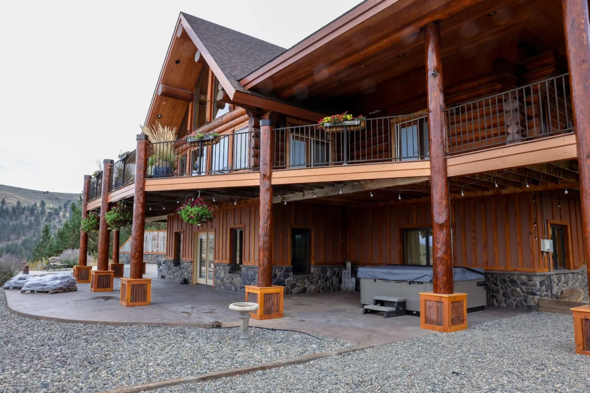 The hero image showcases the grand log exterior of Whispering Pines Lodge, with impressive wooden pillars, warm stone accents, a flower-adorned balcony, and an inviting hot tub set against sweeping views of the surrounding mountains and valley. The setting exudes rustic elegance and is ideal for outdoor relaxation and gatherings under the open sky.