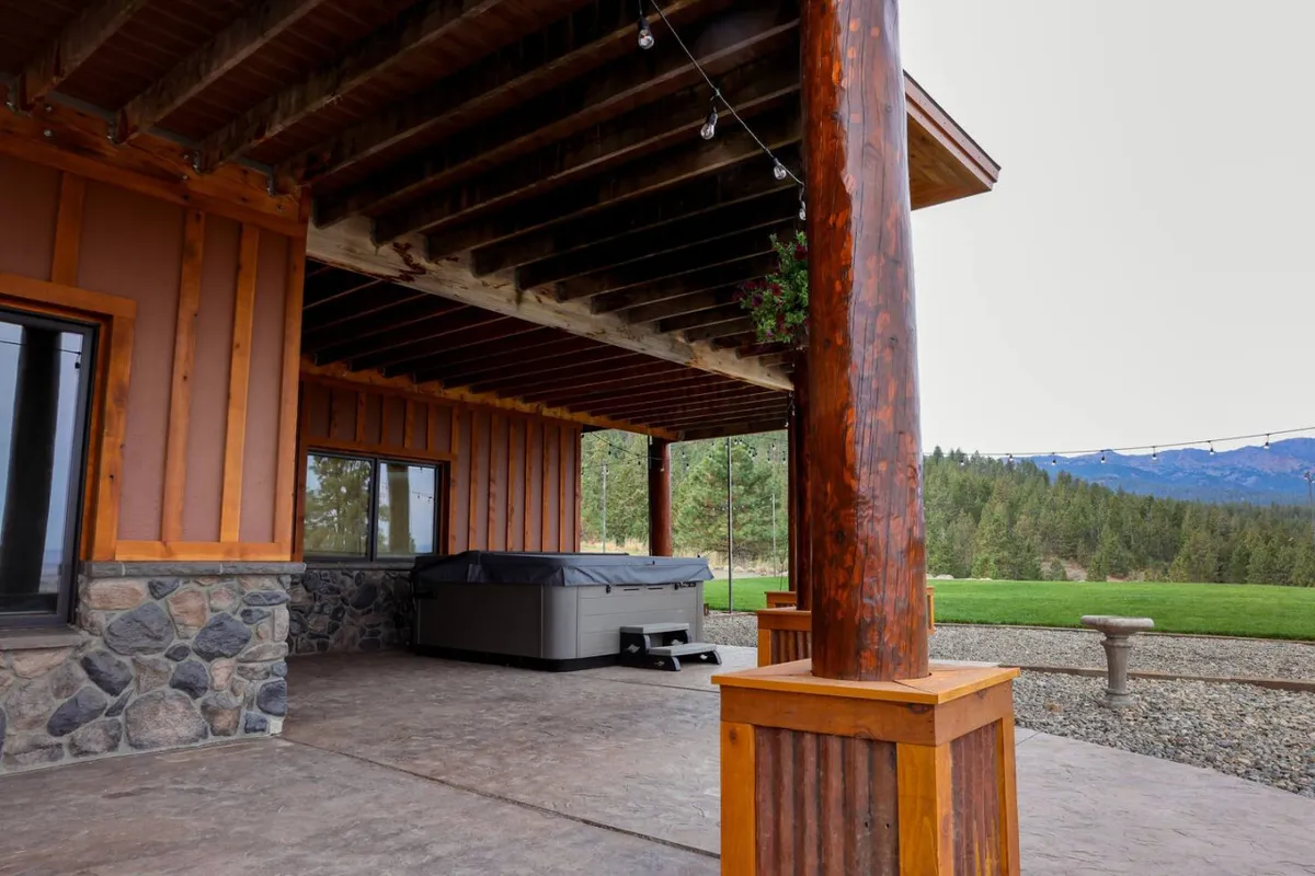 A tranquil outdoor hot tub sits beneath wooden beams and stone walls, surrounded by green landscape and open sky. The view offers a peaceful retreat with distant mountain vistas, ideal for unwinding after a day of exploration.
